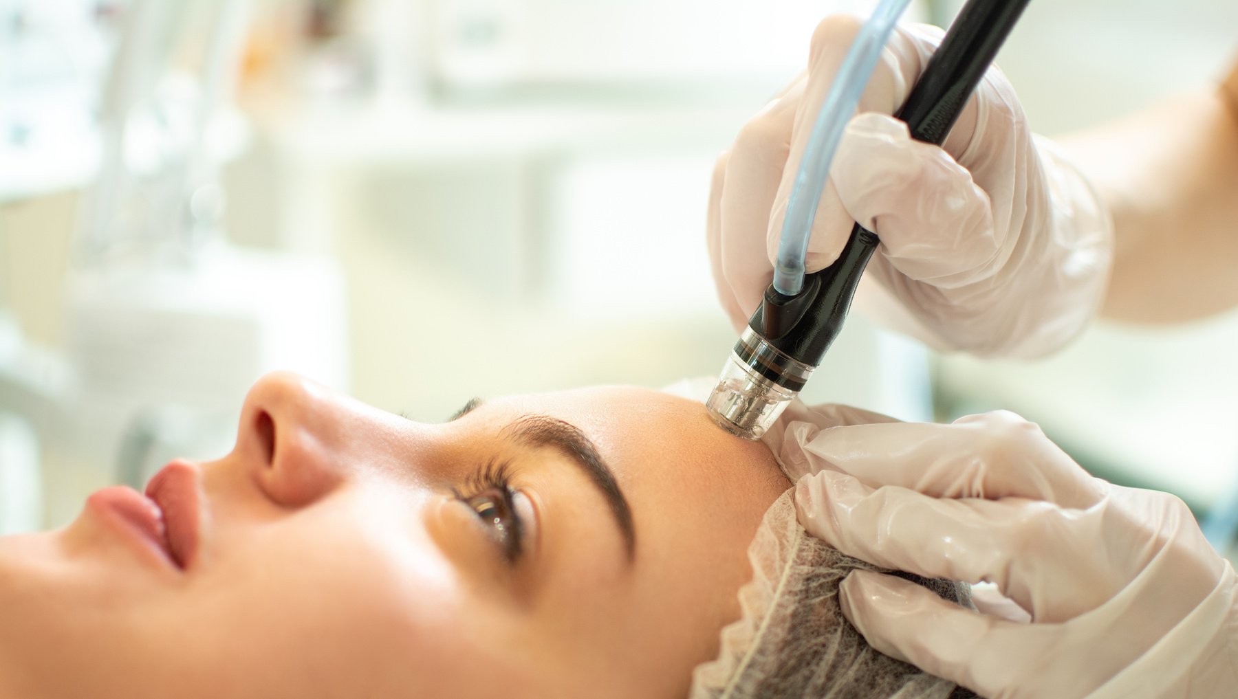 Close up of therapist's hands in protective gloves holding hydrafacial tool and performing anti aging progress to beautiful young woman forehead at beauty salon.